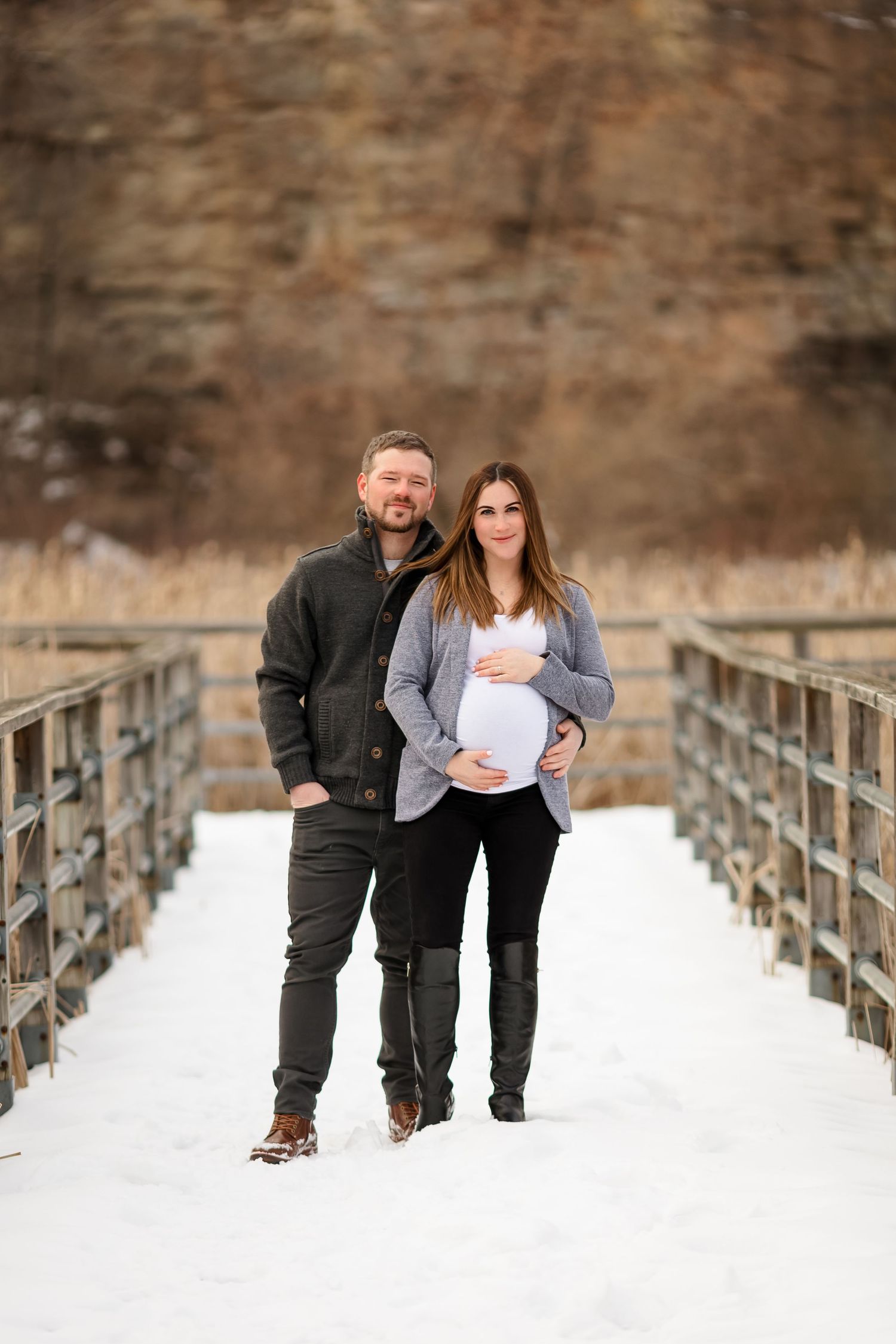 Expectant couple standing together on snow covered bridge during winter photo session.