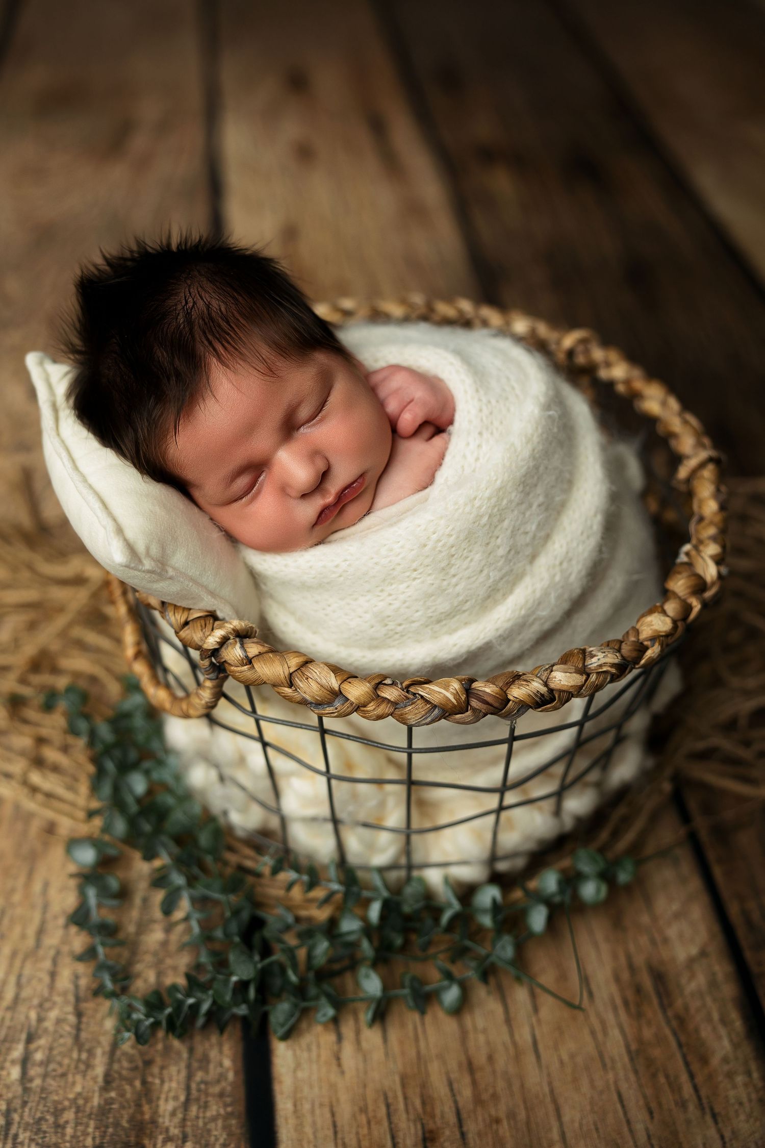 Sweet newborn wrapped in white swaddle sleeping peacefully in woven basket on rustic wooden floor with green garland.