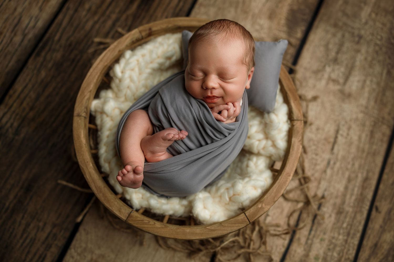 Newborn baby wrapped in gray swaddle sleeping peacefully in a rustic wooden bowl with white fluffy lining.