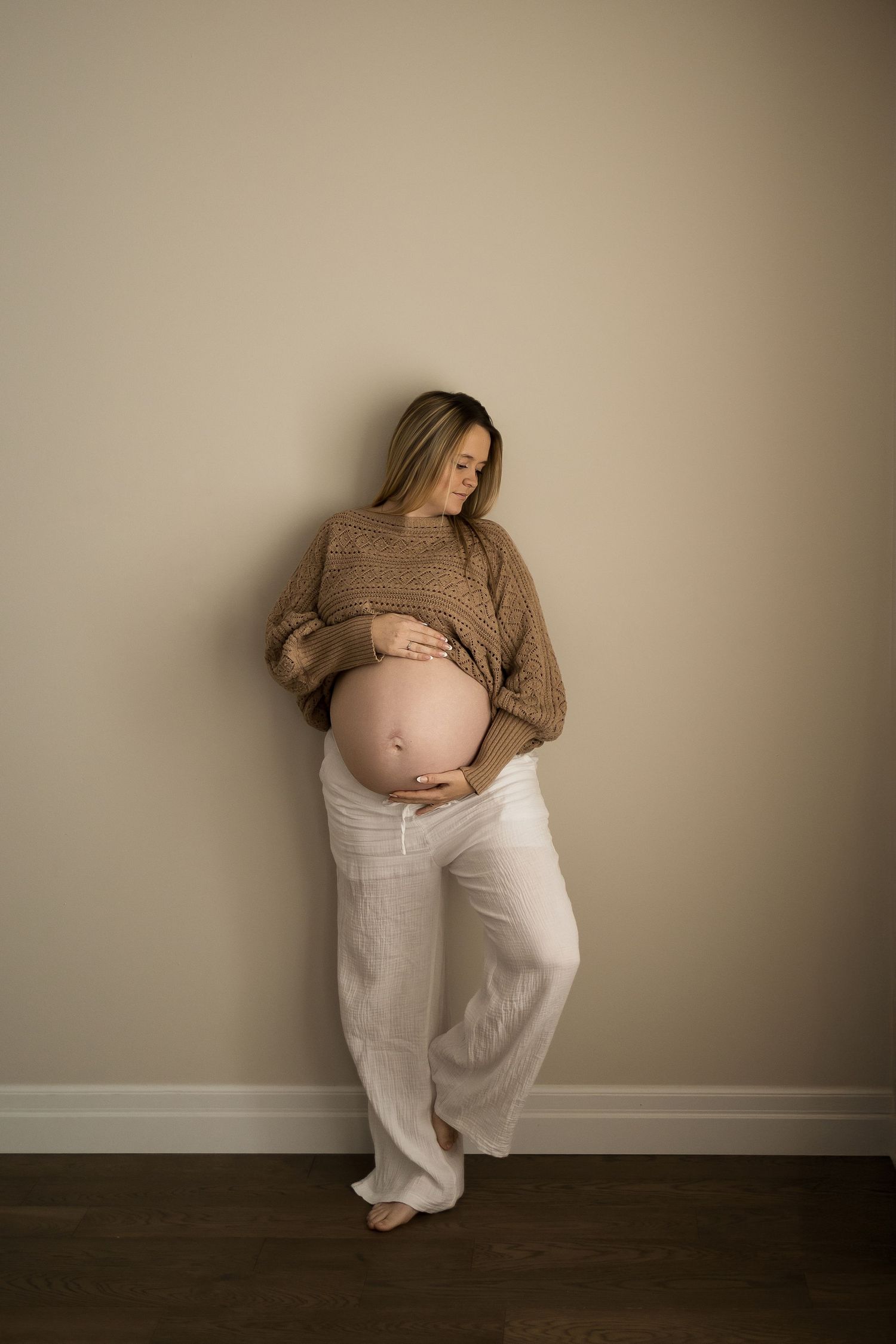 A minimalist maternity photo series against a beige wall showing a person in a cozy sweater and white pants.