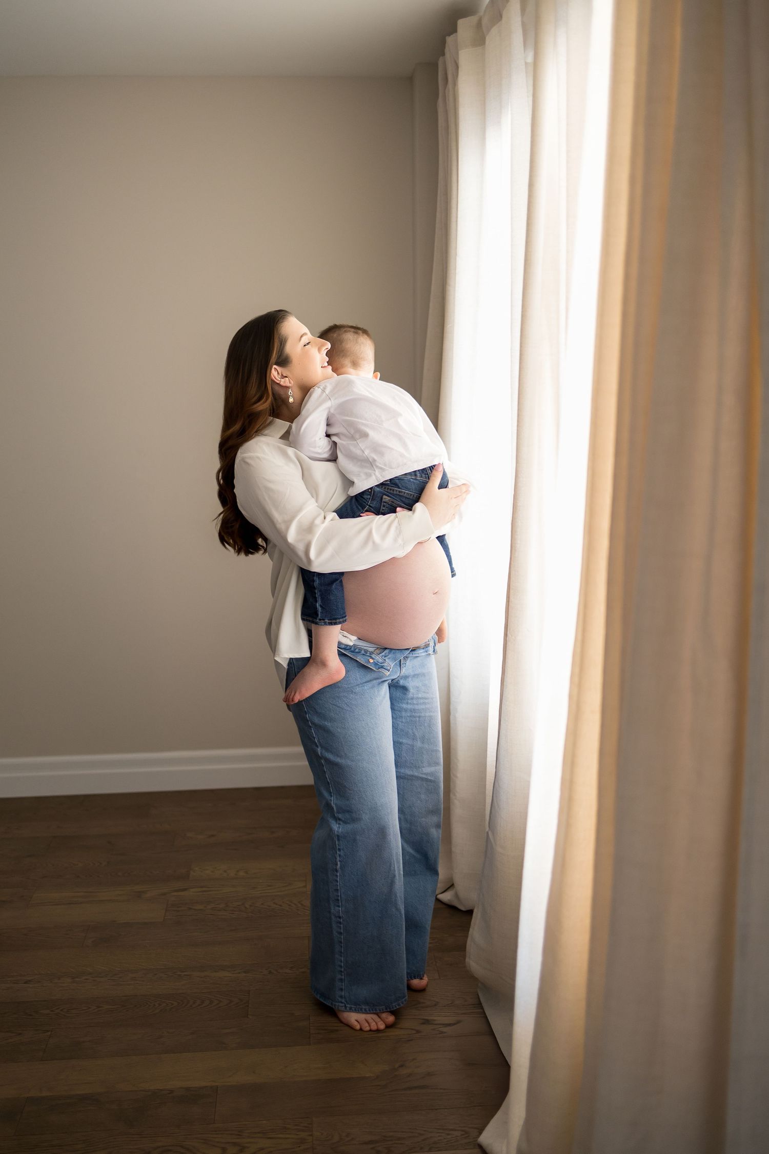 A tender moment by a window as a woman in jeans and white shirt holds a small child in natural lighting.