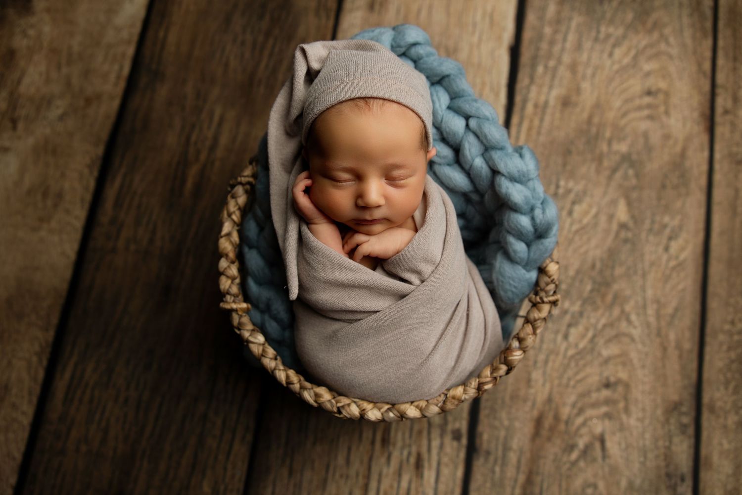 Sleeping newborn in gray wrap with blue knit blanket in round basket.