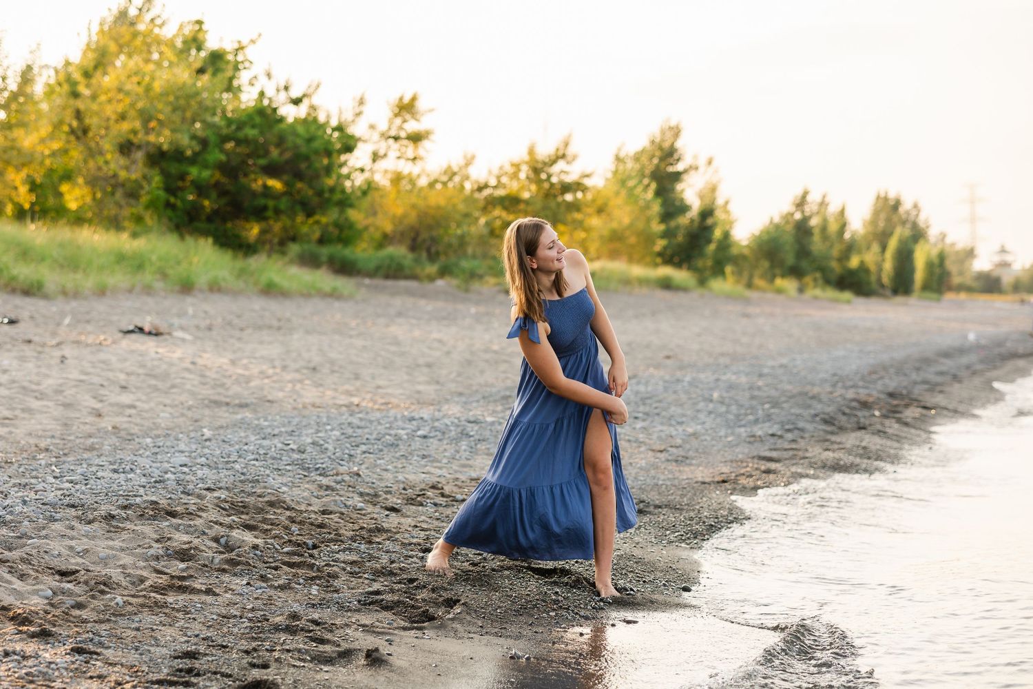 A person in a flowing blue dress twirls at the edge of a beach during golden hour sunset.