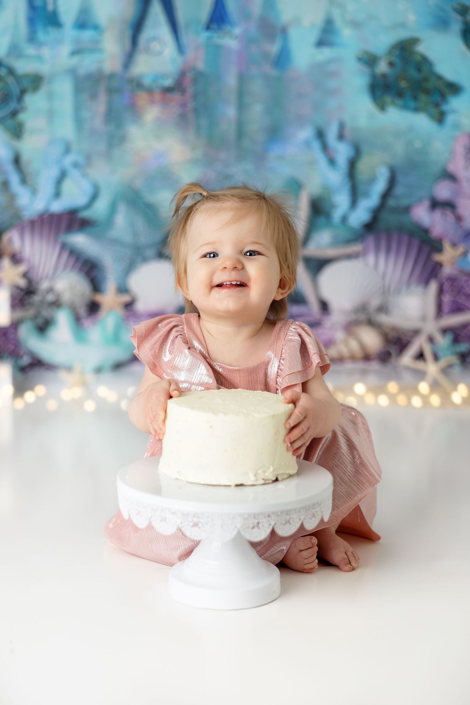 Pink-dressed baby poses with white birthday cake on pedestal against dreamy underwater castle backdrop.