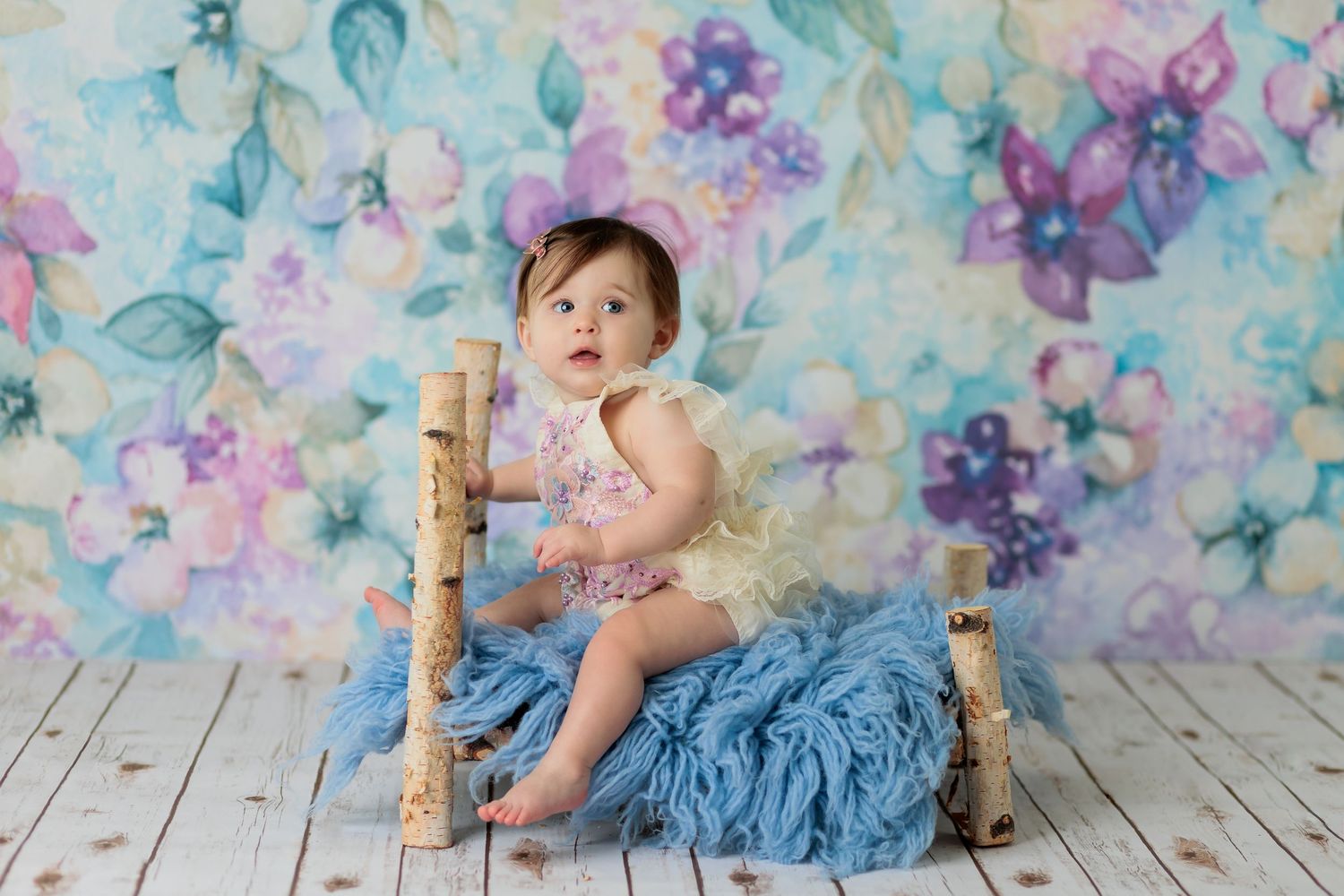 Baby posed on rustic bench with blue fabric against floral backdrop during studio photography session.