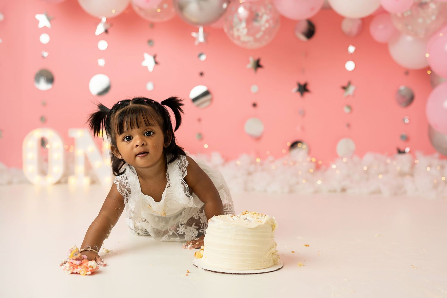 Cake smash photography setup with pink backdrop, silver balloons, and illuminated ONE sign.