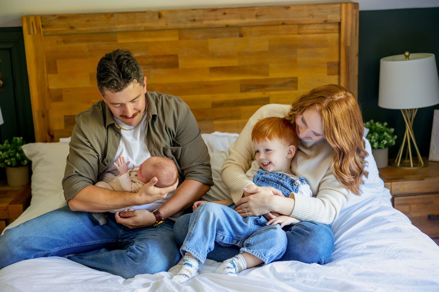 A family shares a cozy moment together on a bed with wooden headboard and warm lighting.