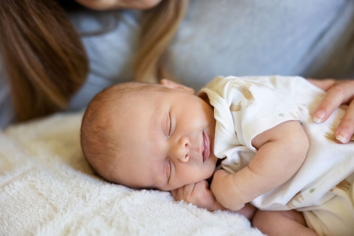 A sleeping newborn baby in white clothing lies on a cream-colored blanket.