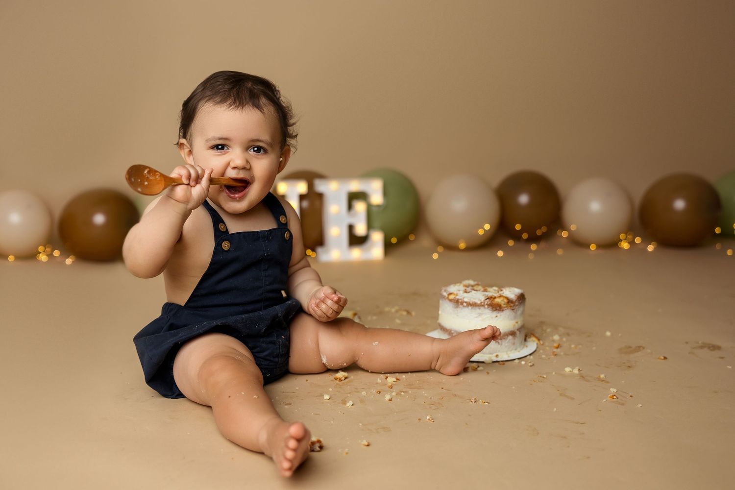Baby in navy blue romper sits on beige backdrop with cake smash setup and brown balloon garland decoration.