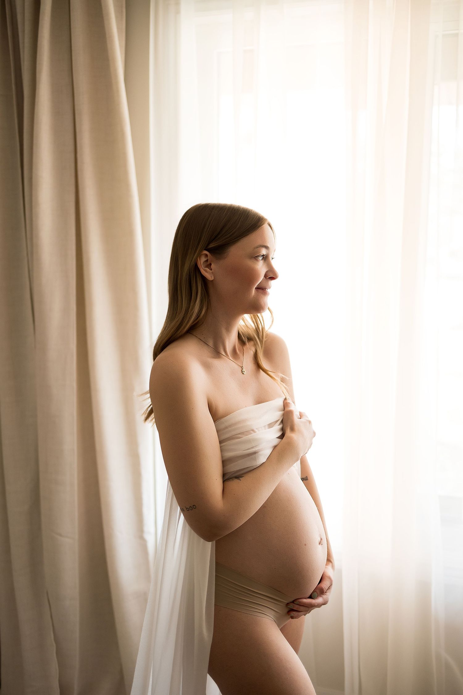 Pregnant woman in beige underwear holding her baby bump near sheer curtains in soft natural light.