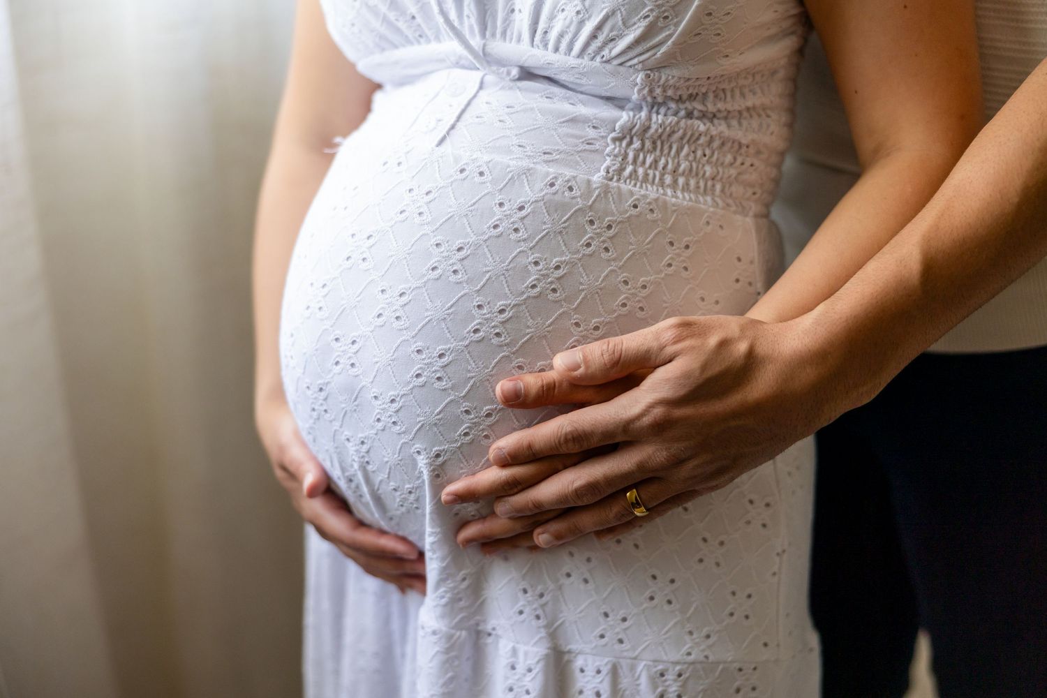 An expecting mother in a white eyelet lace dress gently cradles her belly in soft window light.