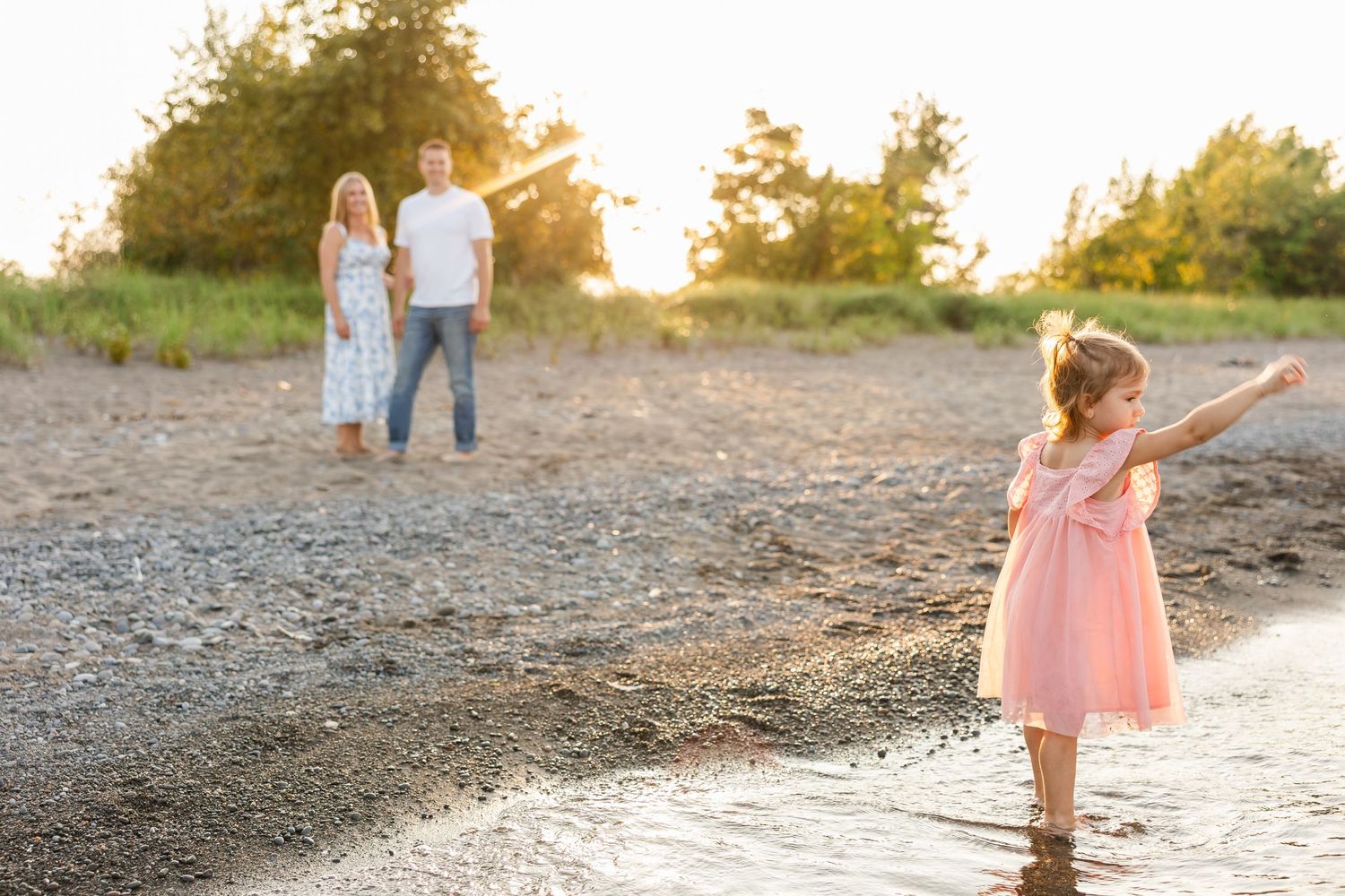 A child in a pink dress plays by the water's edge at sunset while two people stand in the background.