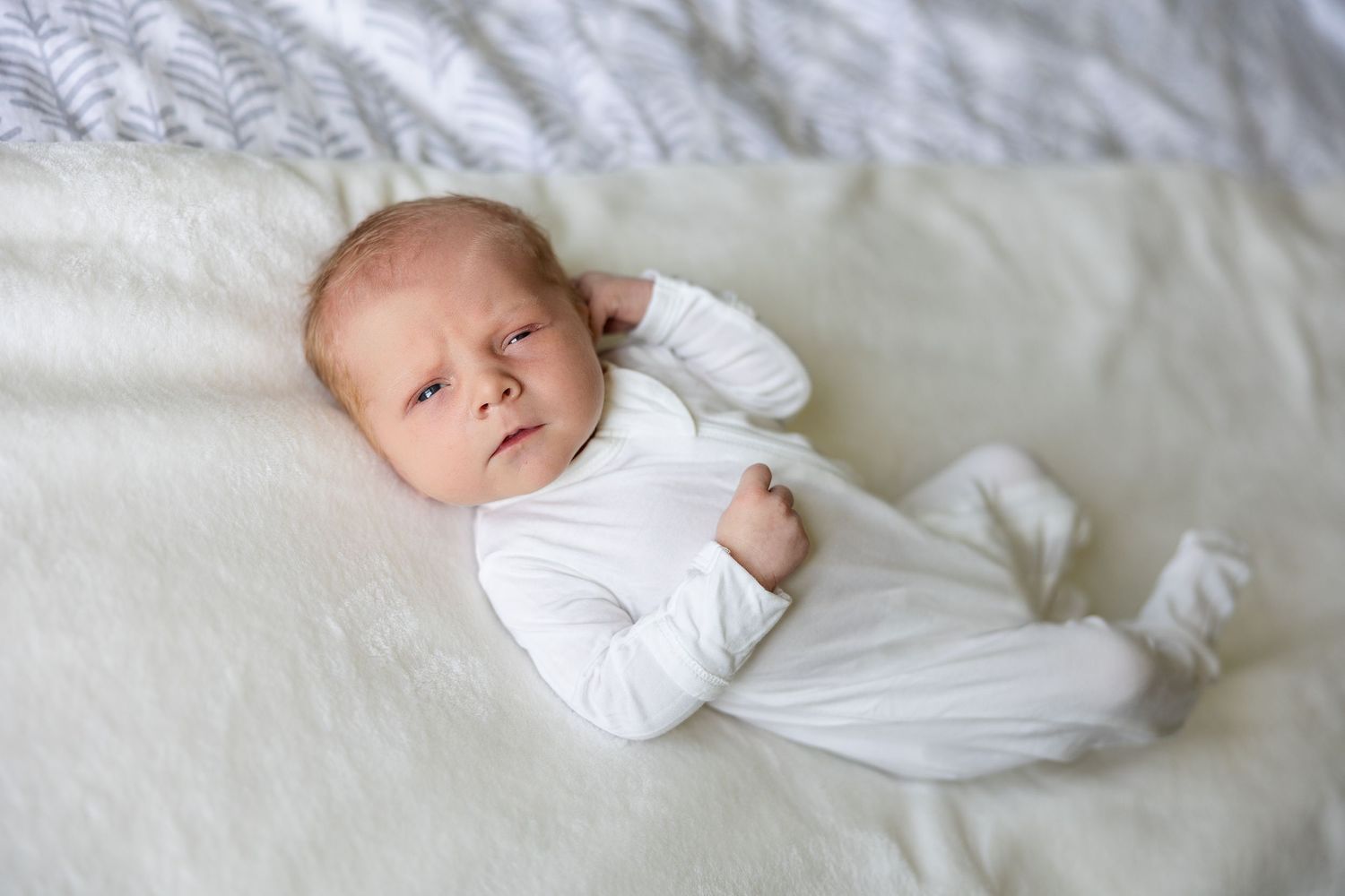 A tiny newborn wearing white sleepwear rests peacefully on soft white bedding with a contemplative expression.