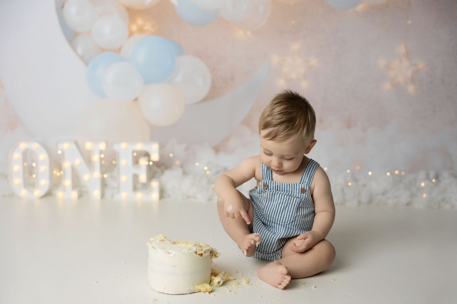 Dreamy first birthday portrait with lit 'ONE' sign, white cake, and soft blue and white balloon decorations.