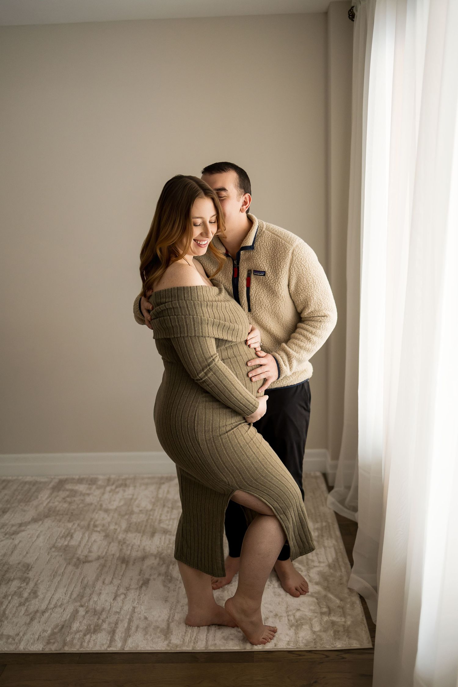 A couple shares an intimate moment in a sunlit room during a maternity photoshoot.