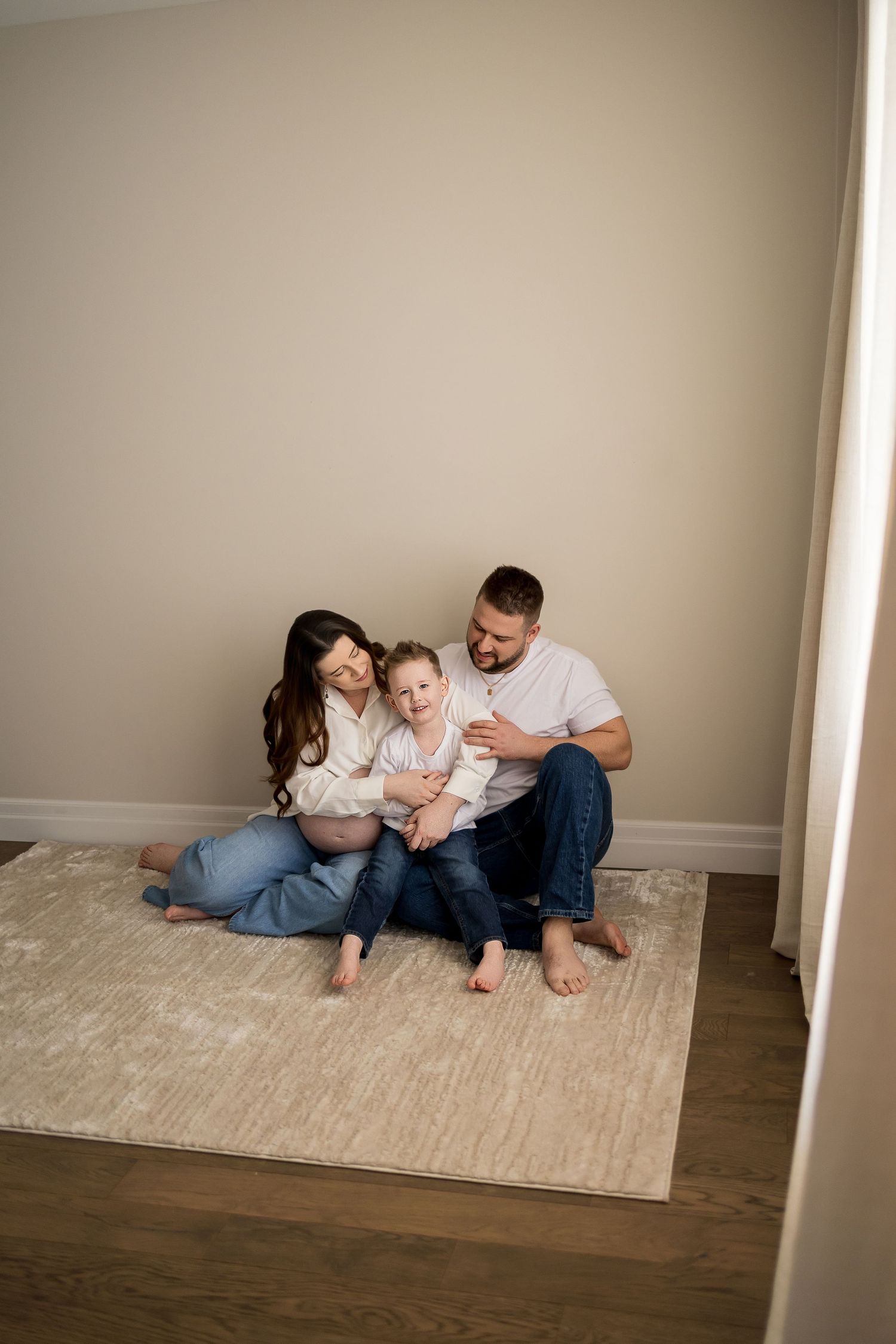 A family sits together on a beige area rug in a minimalist room with hardwood floors and neutral walls.