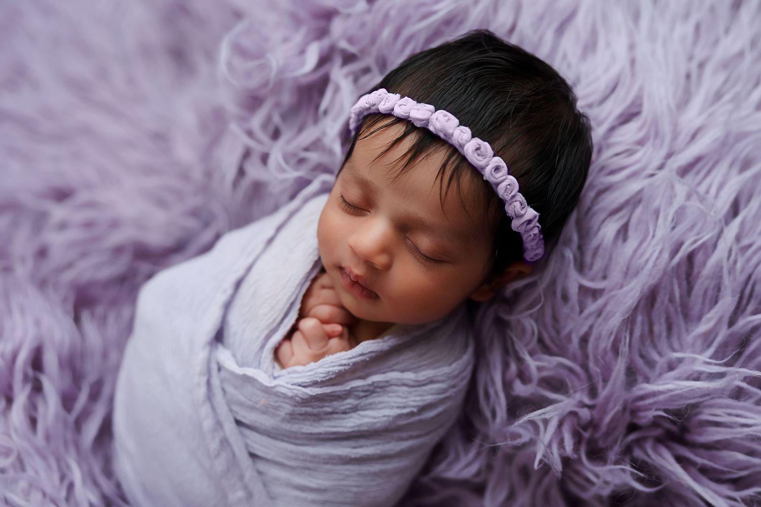 Swaddled newborn resting on lavender fur backdrop with matching headband.