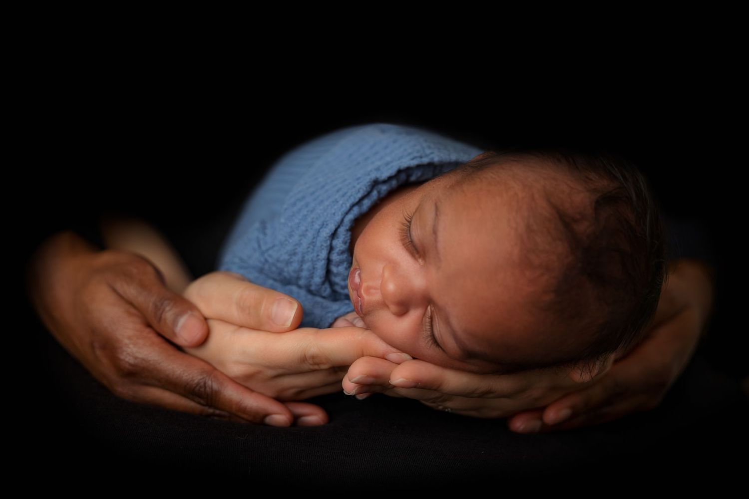 Peaceful newborn baby wrapped in blue blanket resting on hands against dark backdrop.