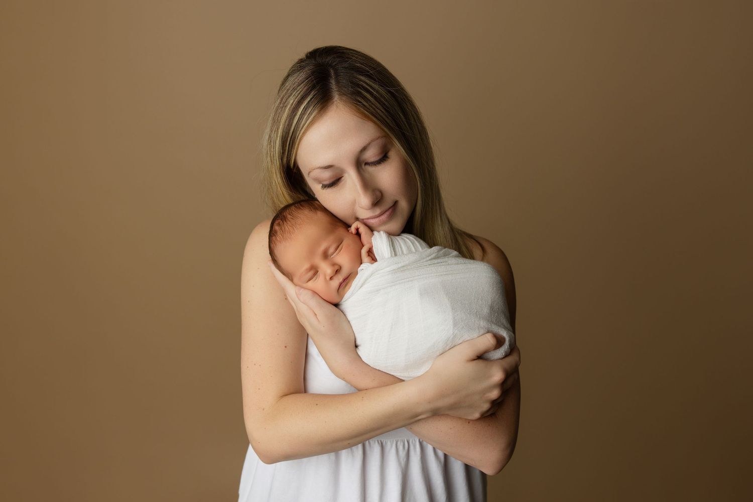 Parent in grey shirt holding sleeping newborn against brown backdrop.