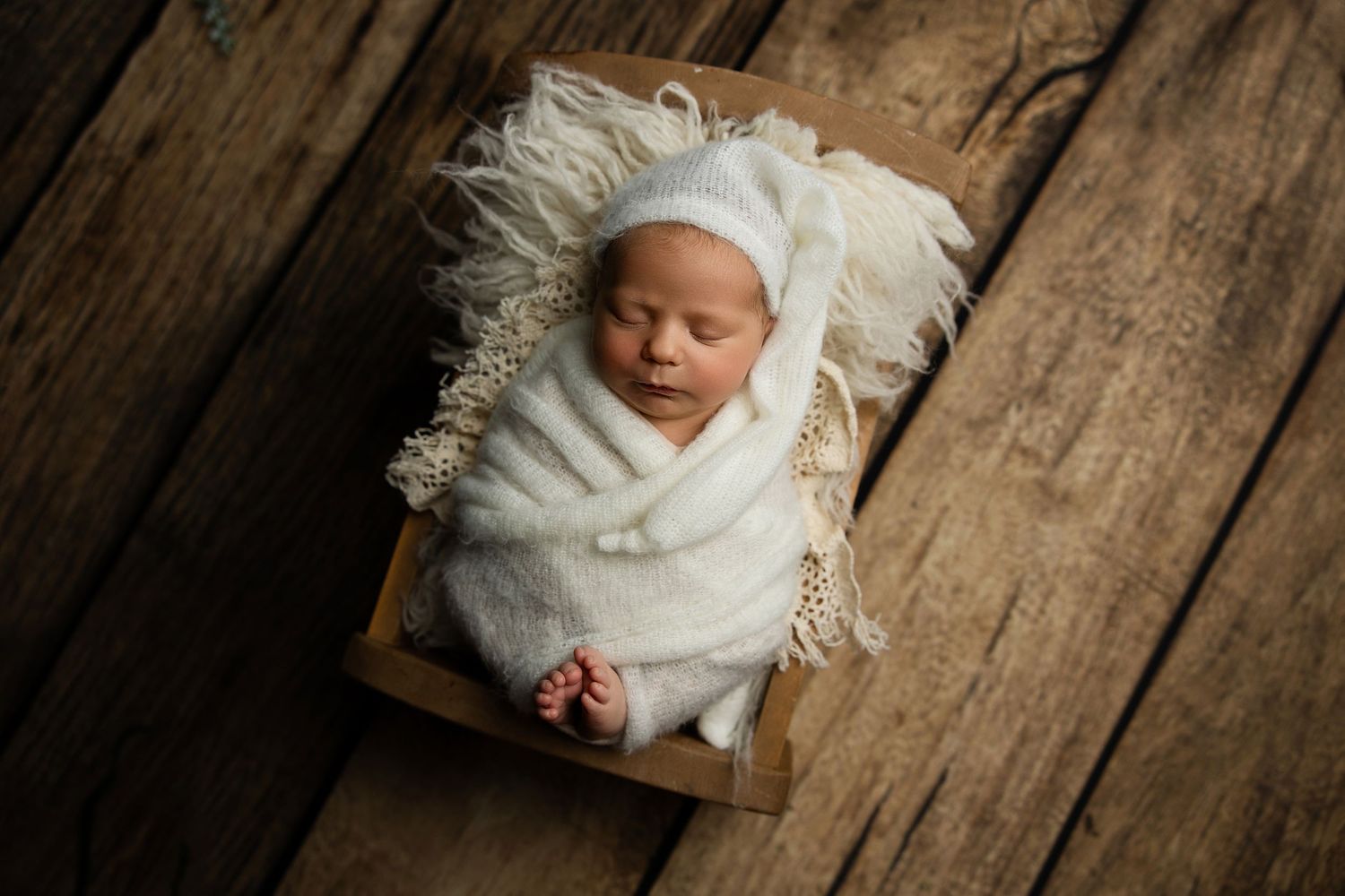 Sleeping infant wrapped in white swaddle blanket on wooden surface with fluffy white texture.