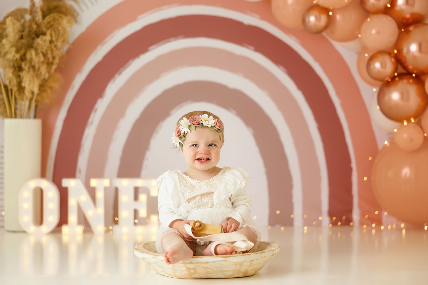 First birthday cake smash photo with rainbow backdrop and pink balloons for milestone celebration.