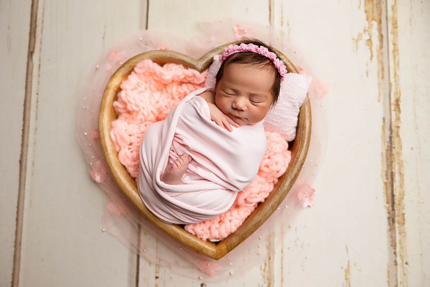 Sleeping baby wrapped in pink swaddle resting in wooden heart-shaped basket with coral padding.