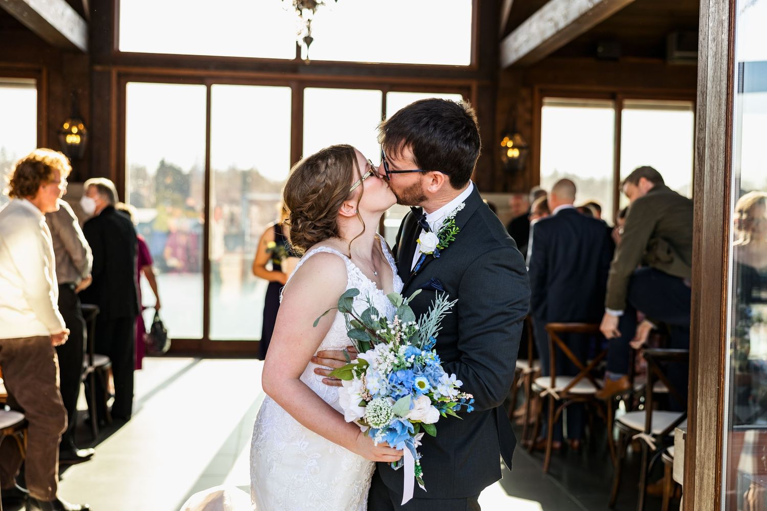 Couple shares romantic kiss during wedding ceremony with blue bouquet and natural light.