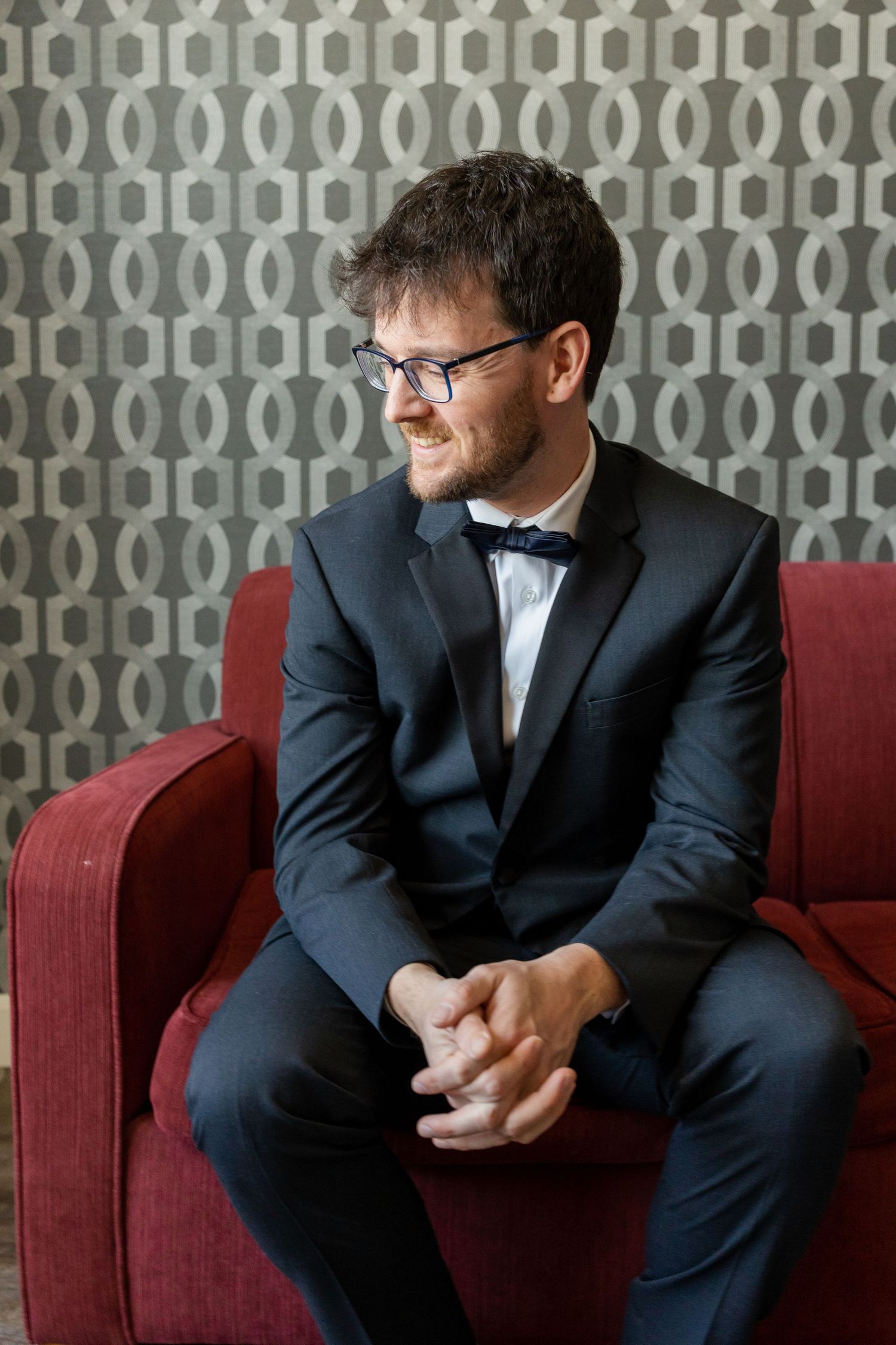 A person in a dark suit and bowtie sits thoughtfully on a red chair against a patterned wall.