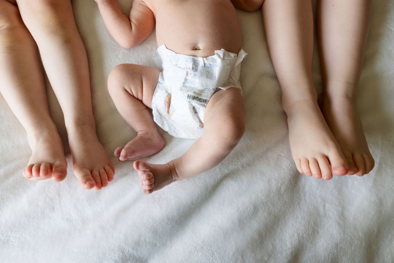 A baby wearing a white diaper lies between adult feet on a soft white blanket.