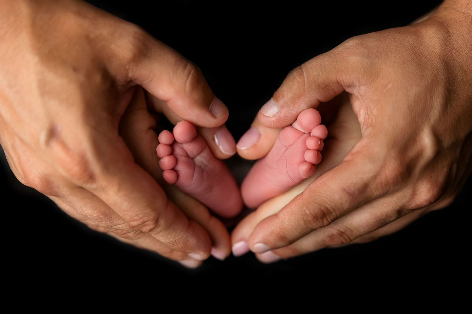 Adult hands form a heart shape around tiny newborn baby feet against a black background.