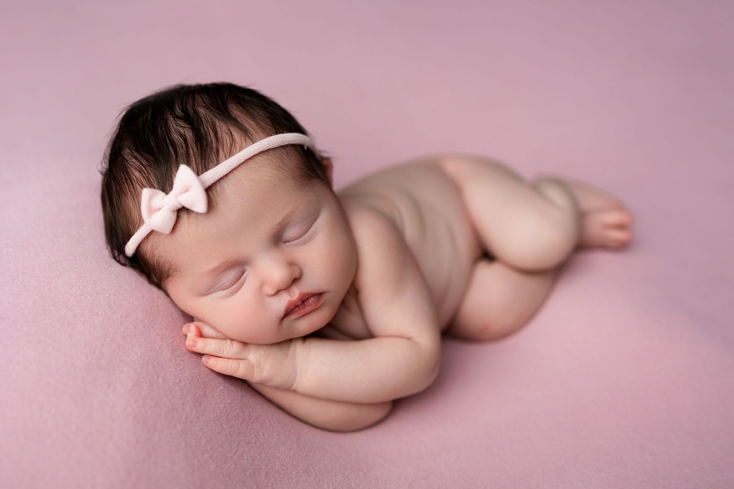 Sleeping newborn with white bow headband on pink background.