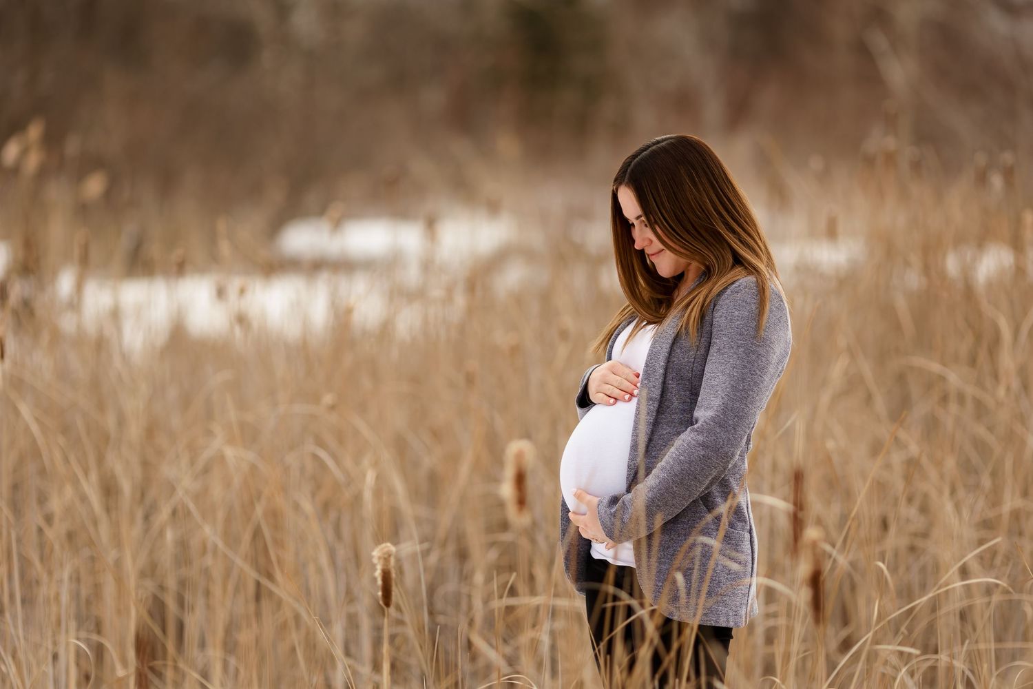 Pregnant woman in gray cardigan stands in winter field during golden hour maternity session.