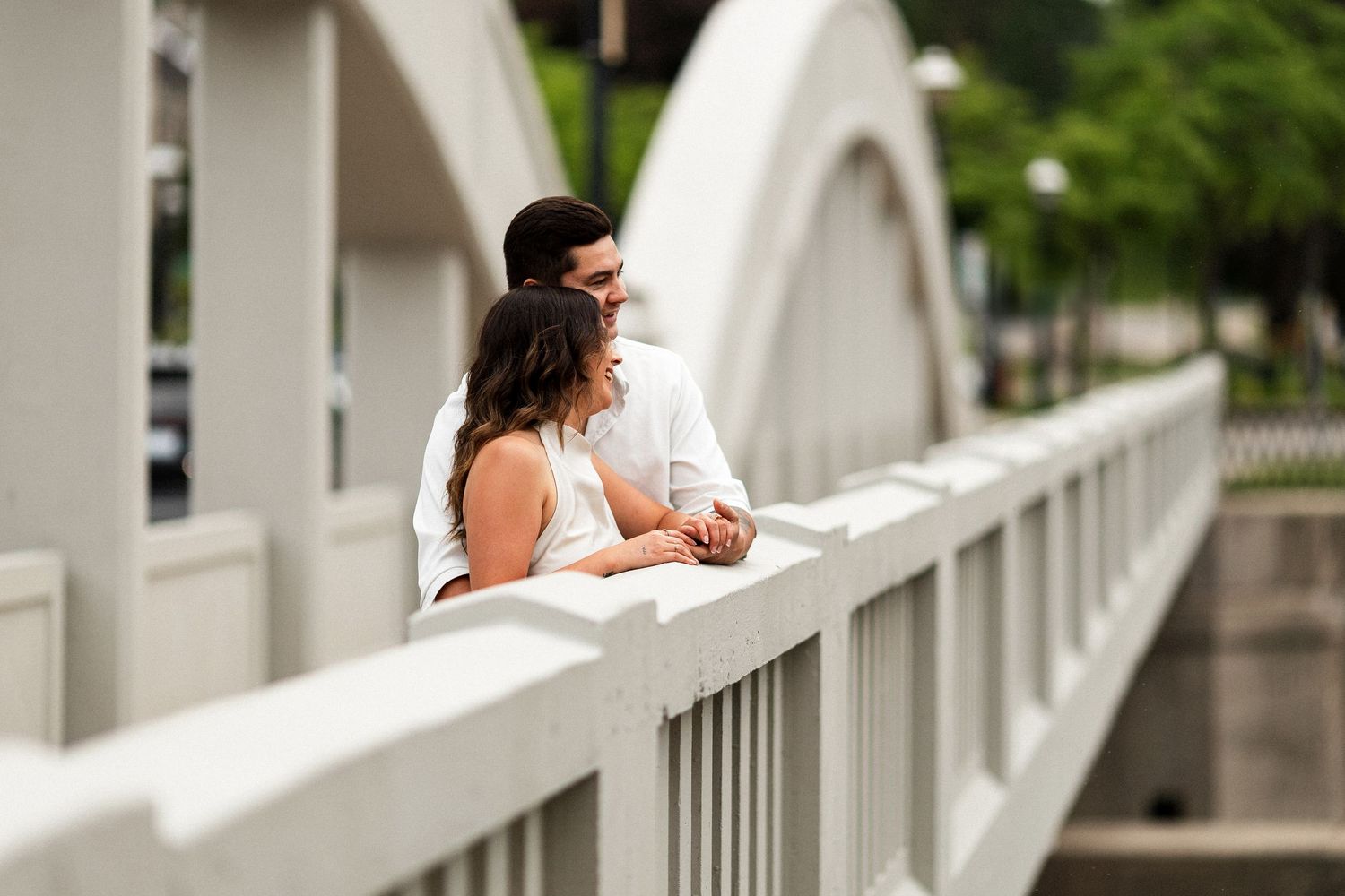 A couple in white attire shares a romantic moment on a decorative white bridge surrounded by lush greenery.
