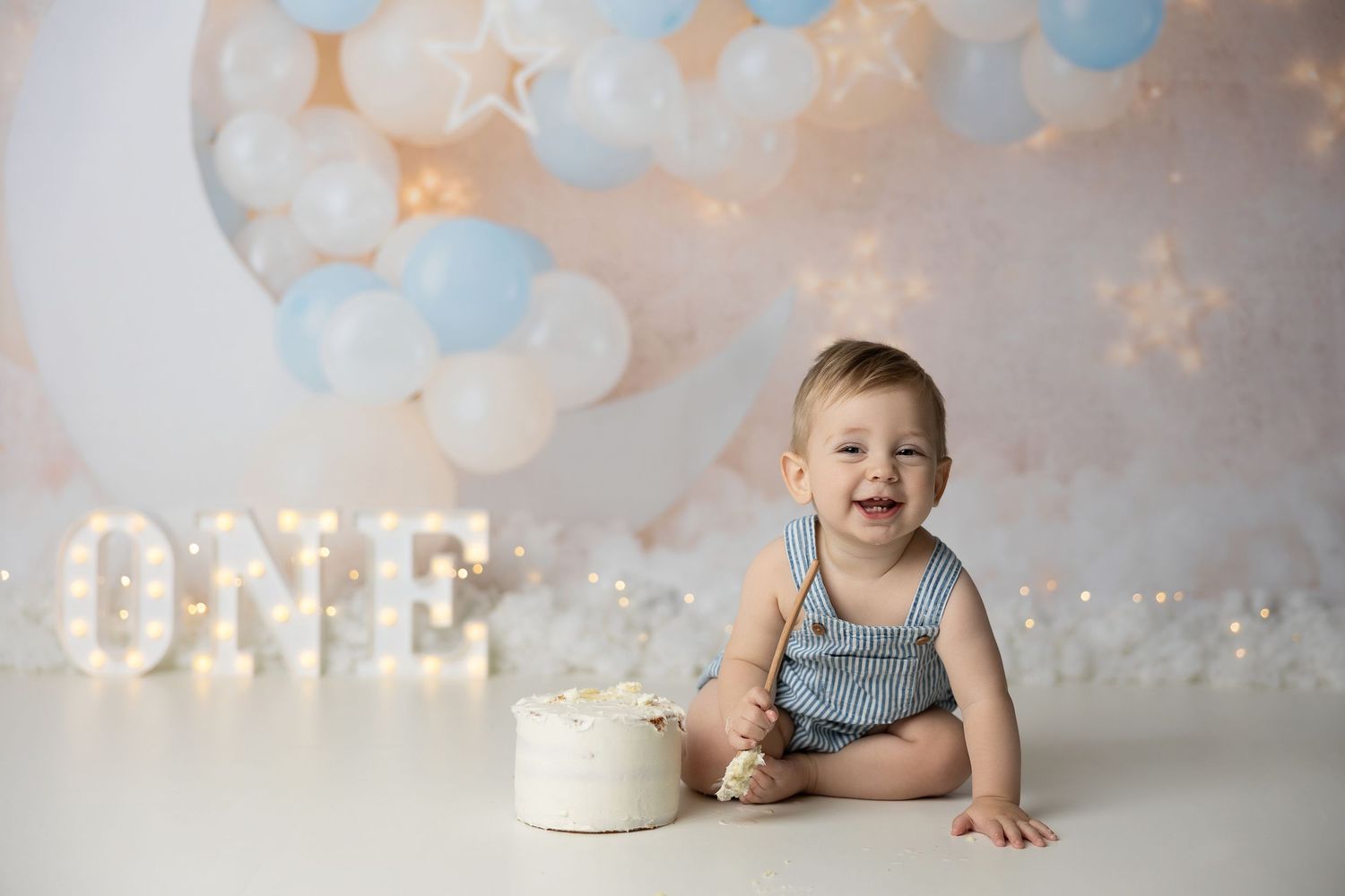Happy cake smash session with illuminated 'ONE' sign and blue and white balloon decor against neutral backdrop.
