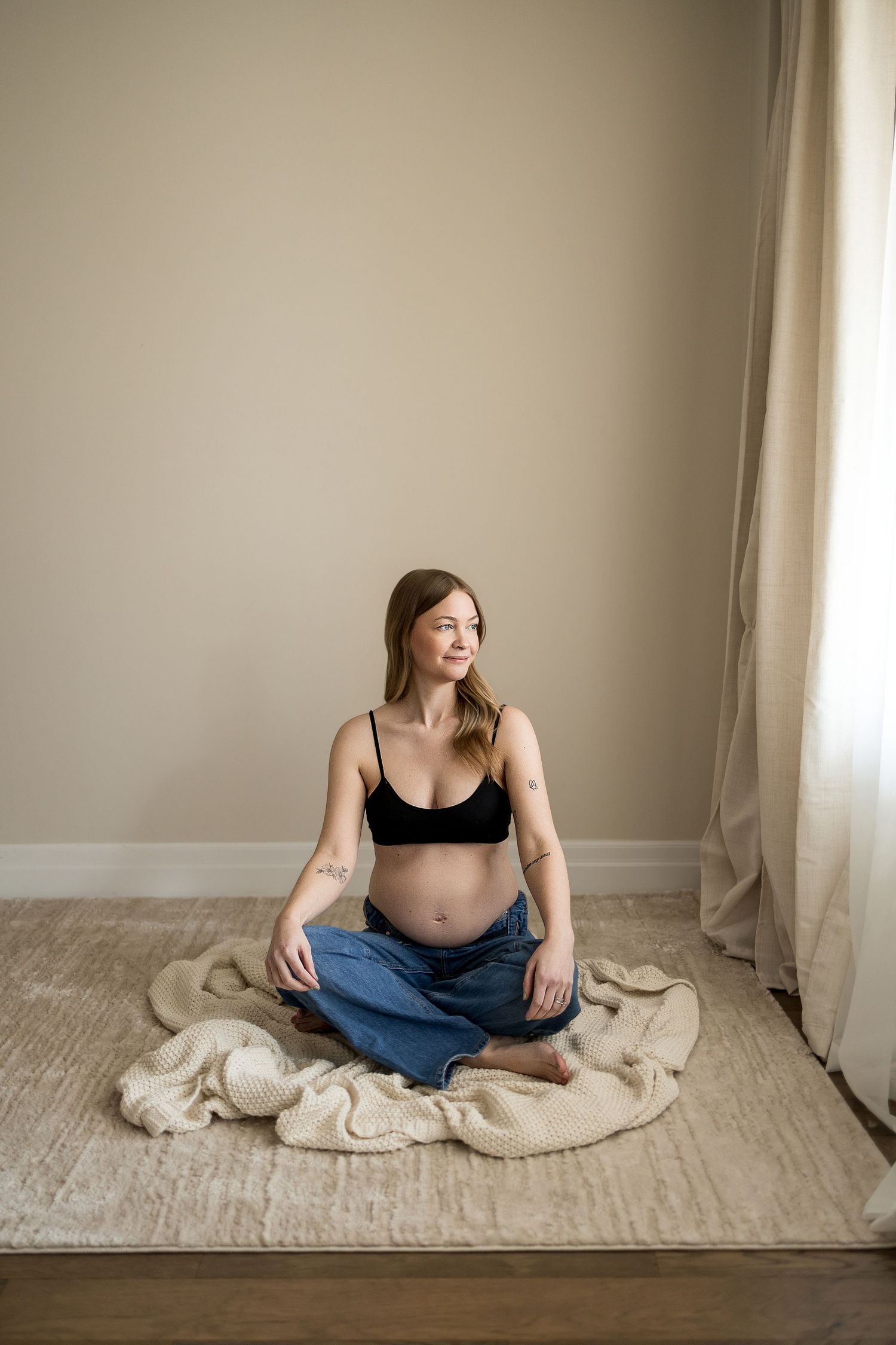 Pregnant woman sitting cross-legged on floor in black crop top and jeans, cradling baby bump in serene home setting.