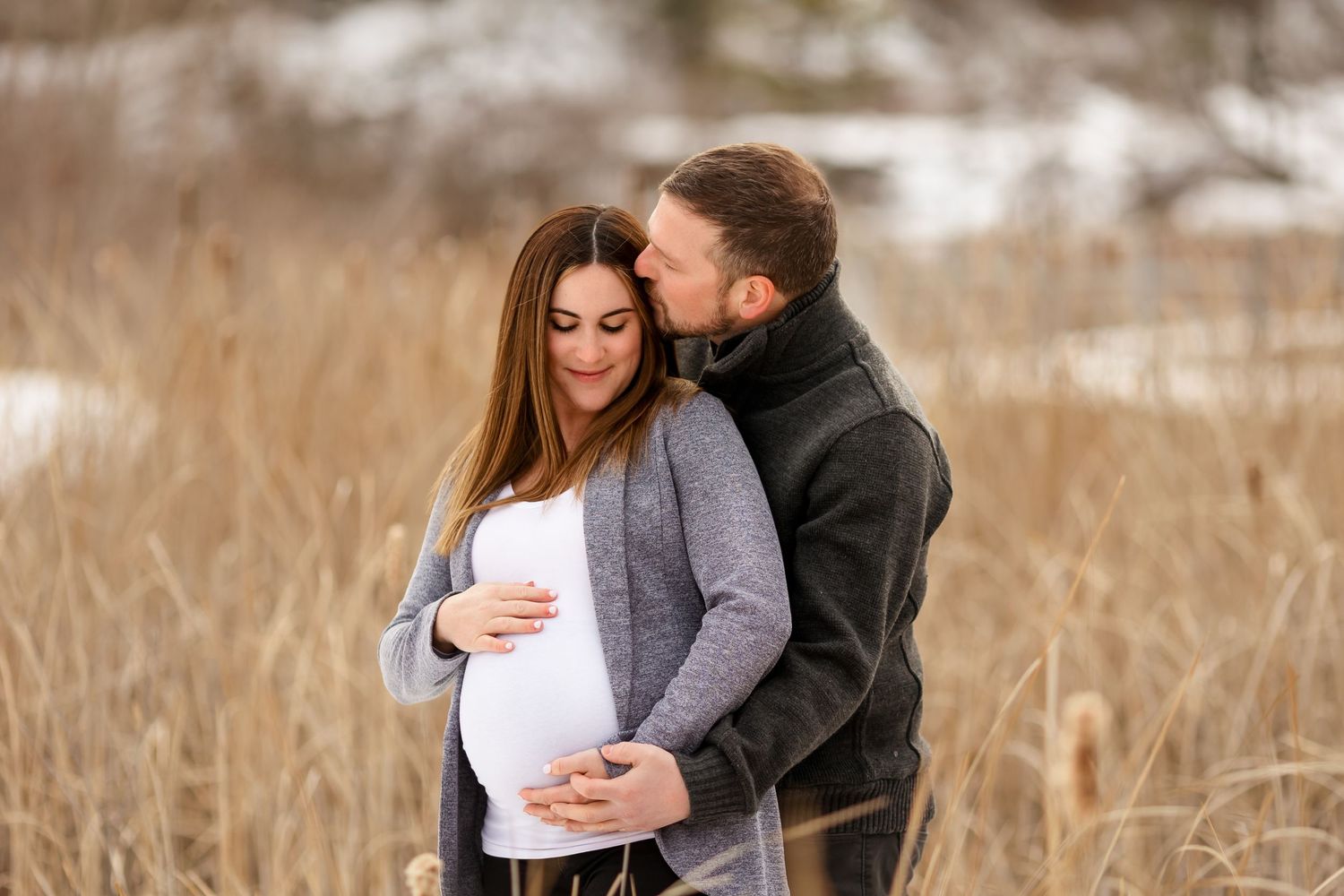 Intimate maternity moment between couple embracing in snowy winter field.