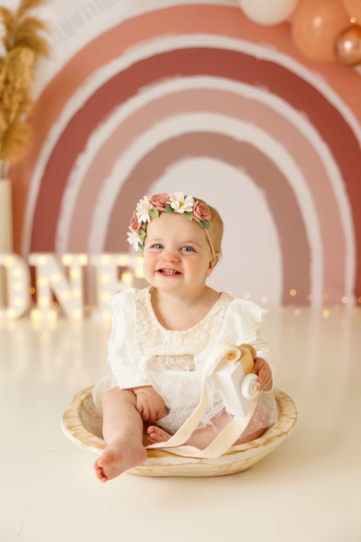 Baby in white dress sitting in wooden bowl against pink rainbow backdrop with balloons.