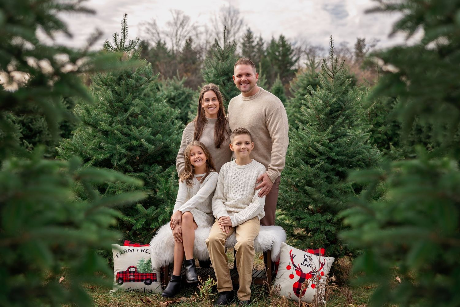 Family holiday photo amongst evergreen Christmas trees with wrapped presents on the ground.