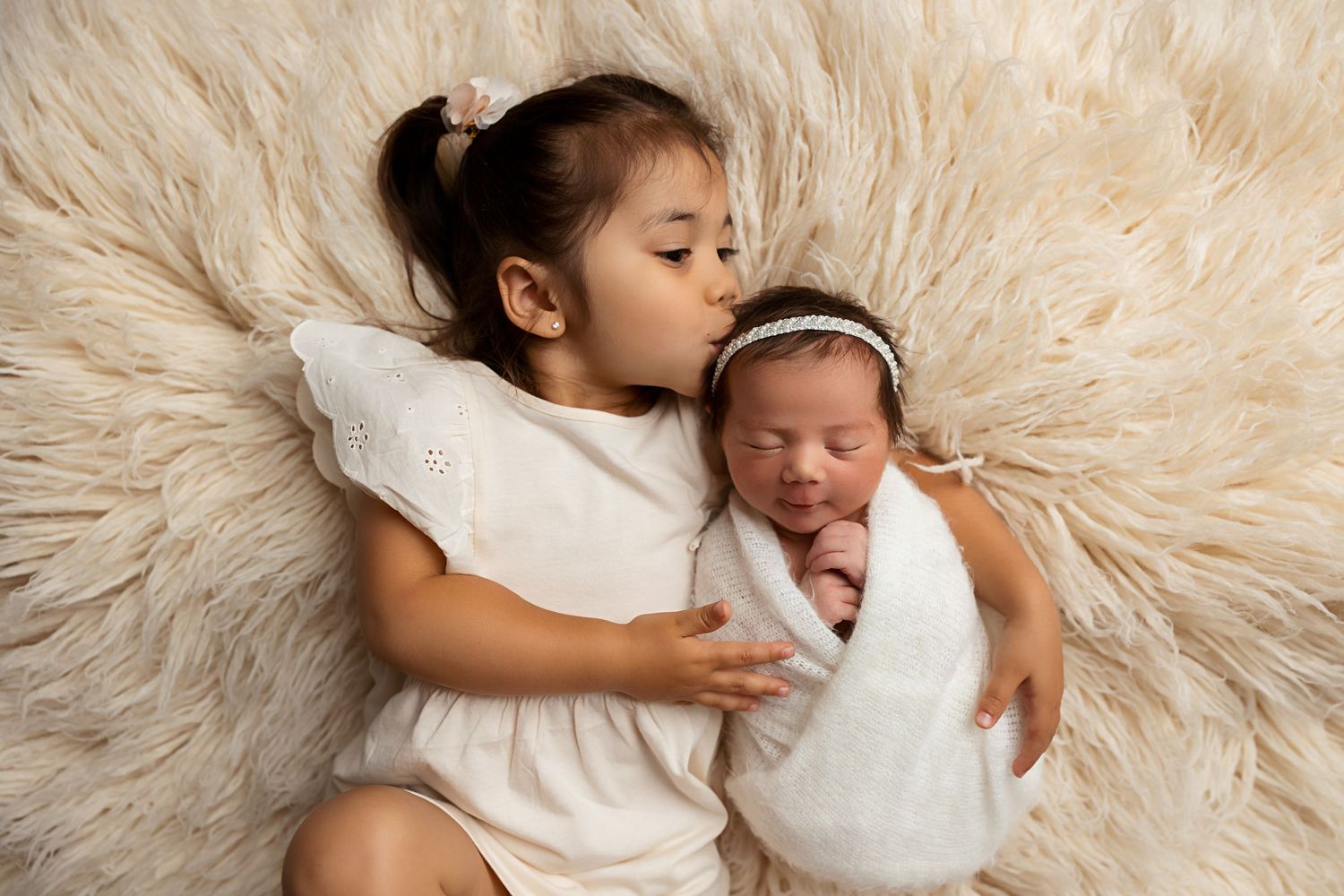 Two siblings lying on fluffy cream rug with newborn baby wrapped in white.