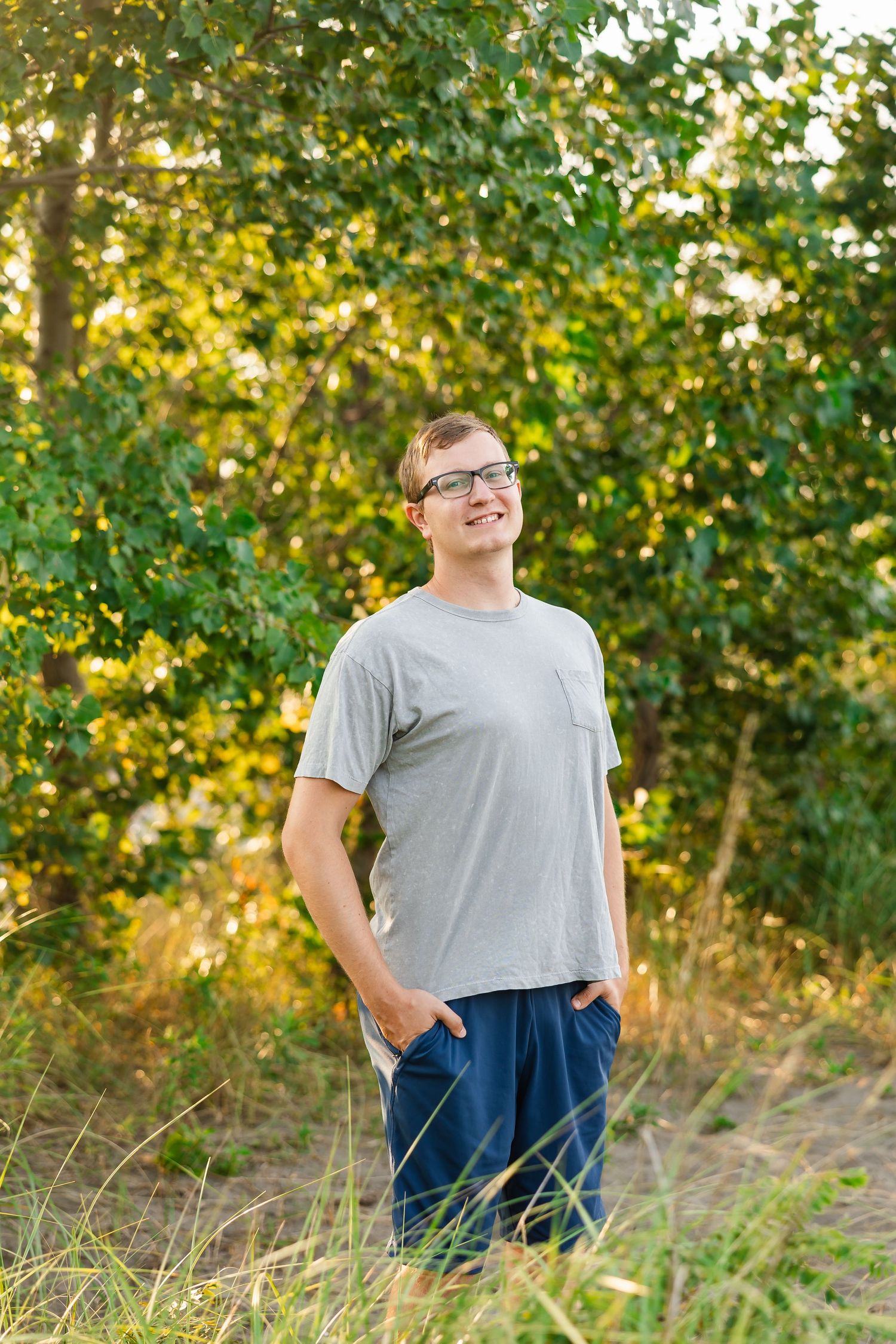 A person in a gray t-shirt and jeans stands outdoors smiling with hands in pockets against a backdrop of sunlit trees.