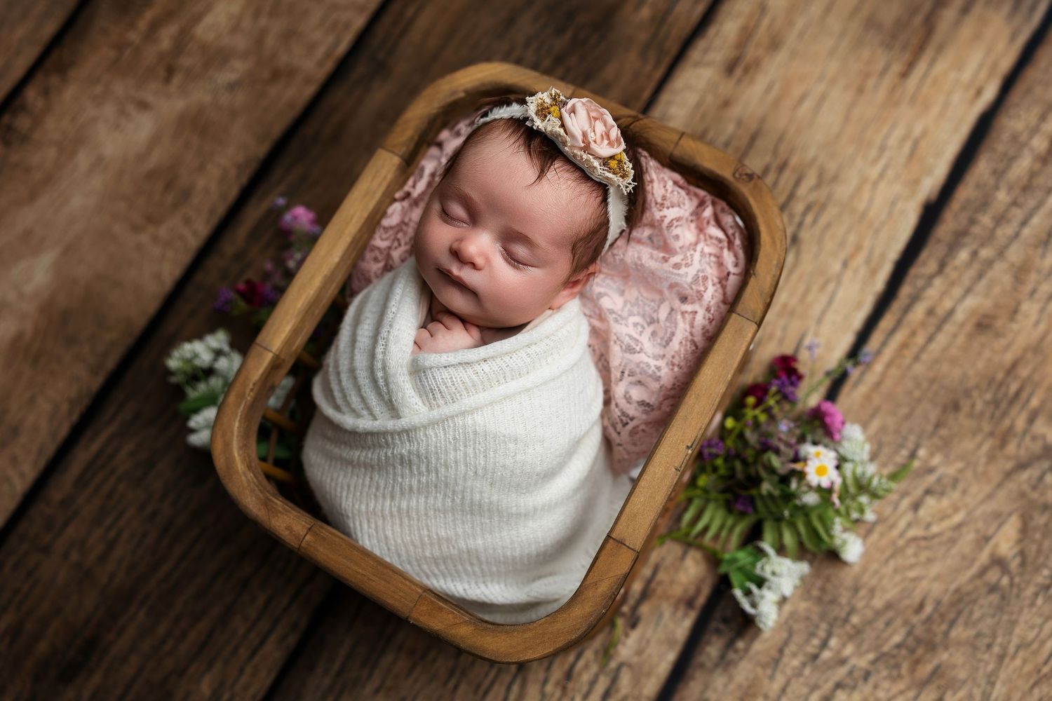 Sleeping newborn wrapped in white swaddle lying in wooden bowl with pink headband and floral decor on wooden floor.