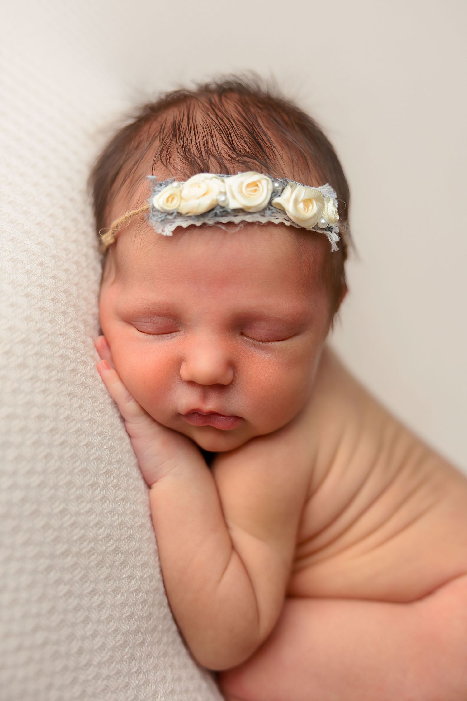 Sleeping newborn with delicate white floral headband resting peacefully on textured white blanket.