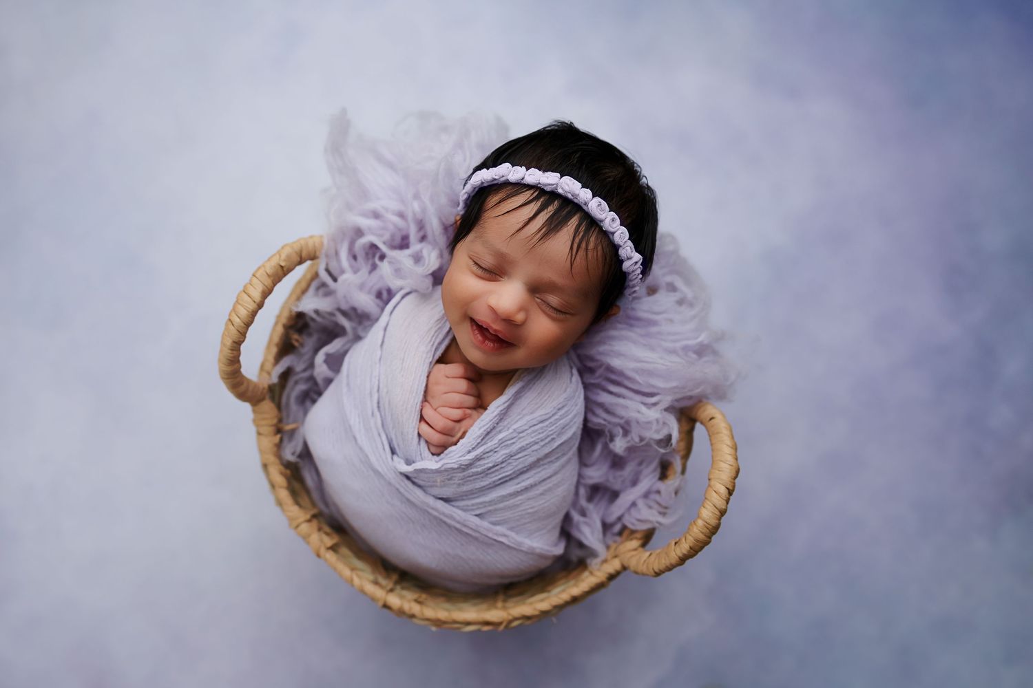 Sweet newborn wrapped in gray swaddle sleeping peacefully in woven basket with purple fabric.