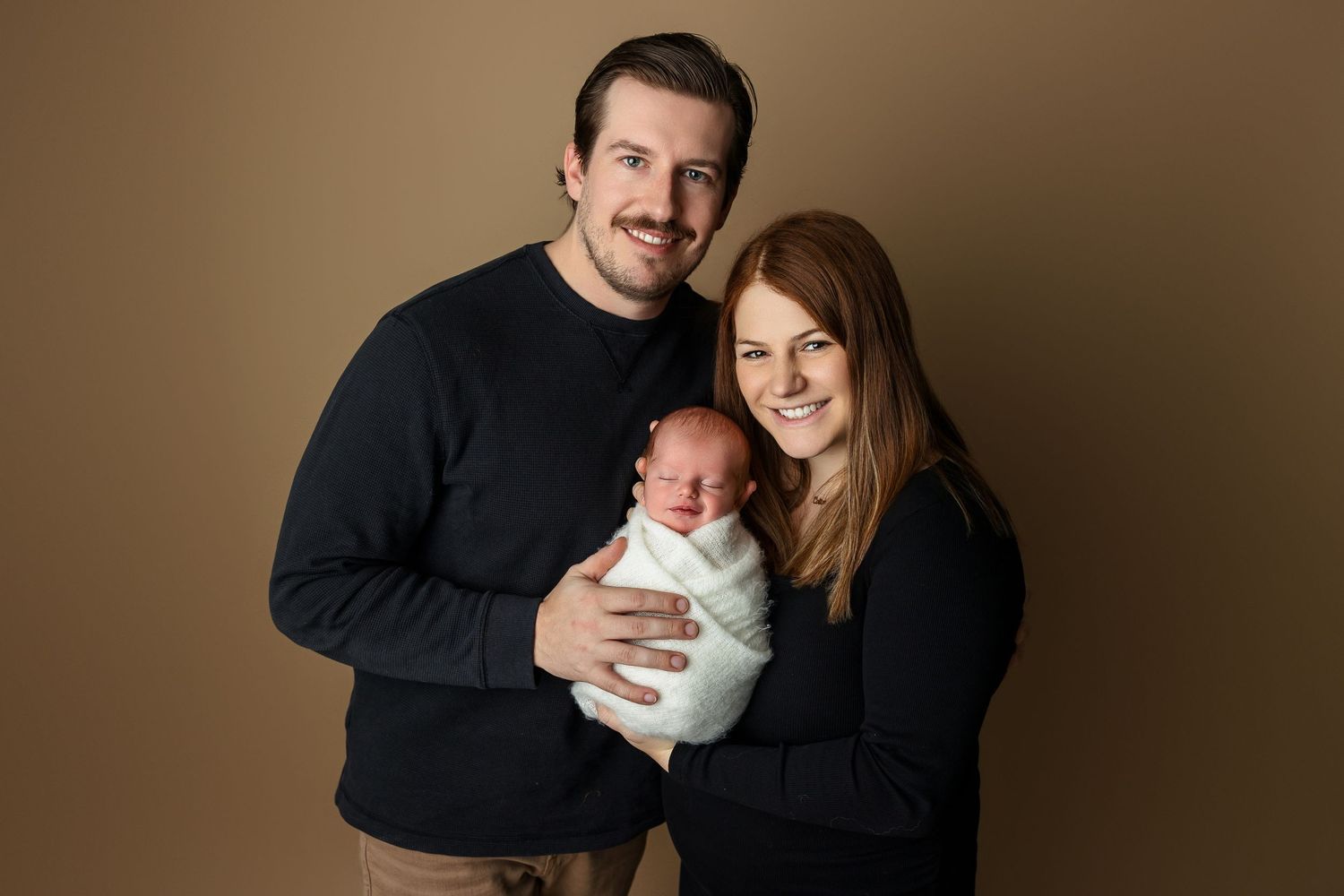 Happy couple in matching dark sweaters holds swaddled newborn against brown background.
