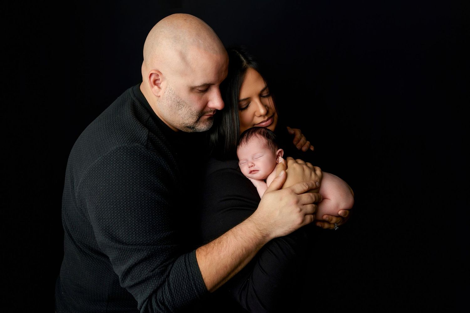 Parents sharing intimate moment with newborn baby against black background in studio setting.