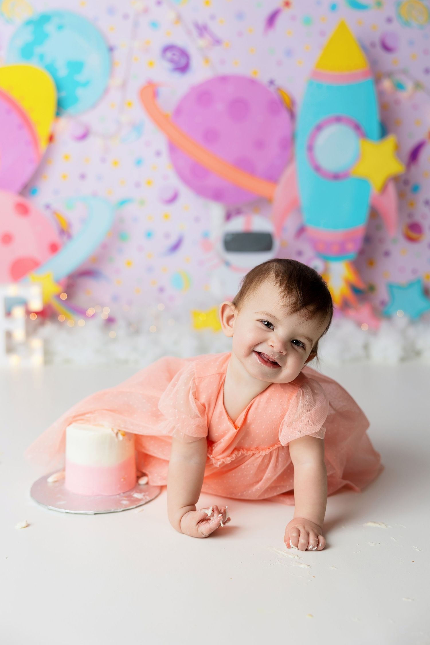 Pink-dressed baby poses near a cake against colorful space-themed backdrop with rockets and planets for birthday shoot.