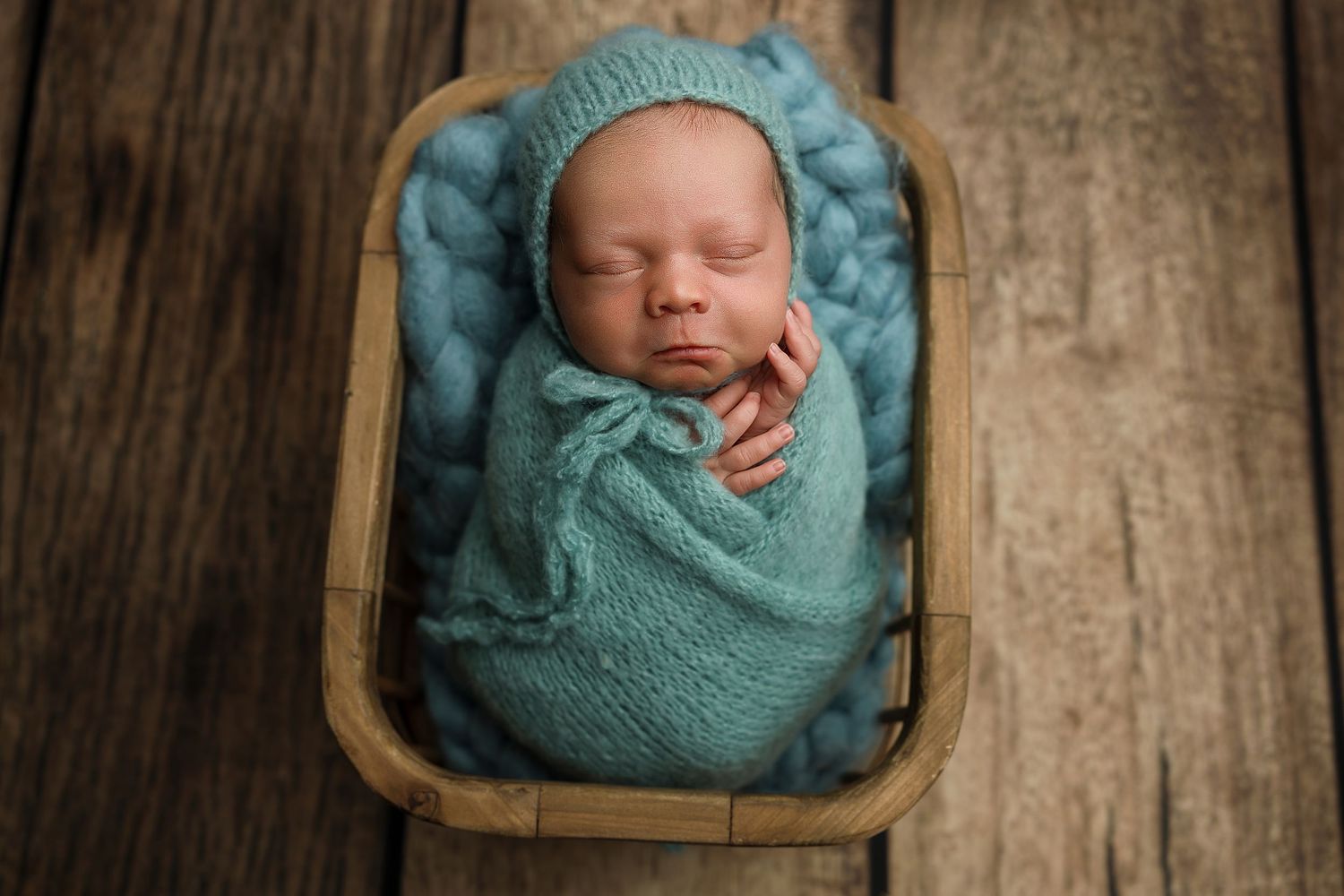 Newborn baby swaddled in teal wrap with knit bonnet, sleeping peacefully in a wooden basket on rustic wood.