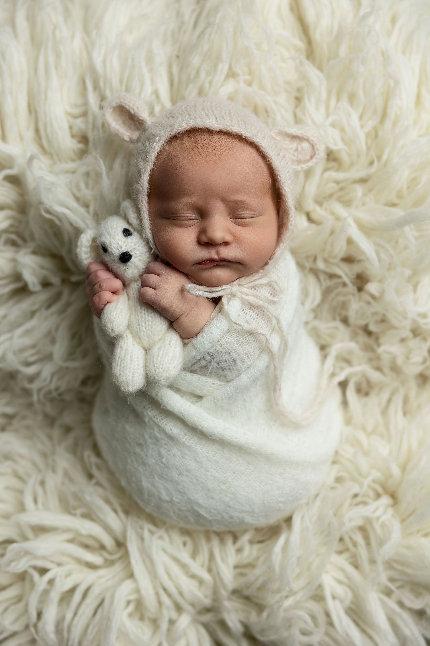 Sleeping newborn swaddled in white on fluffy cream fur blanket with small stuffed bunny toy.