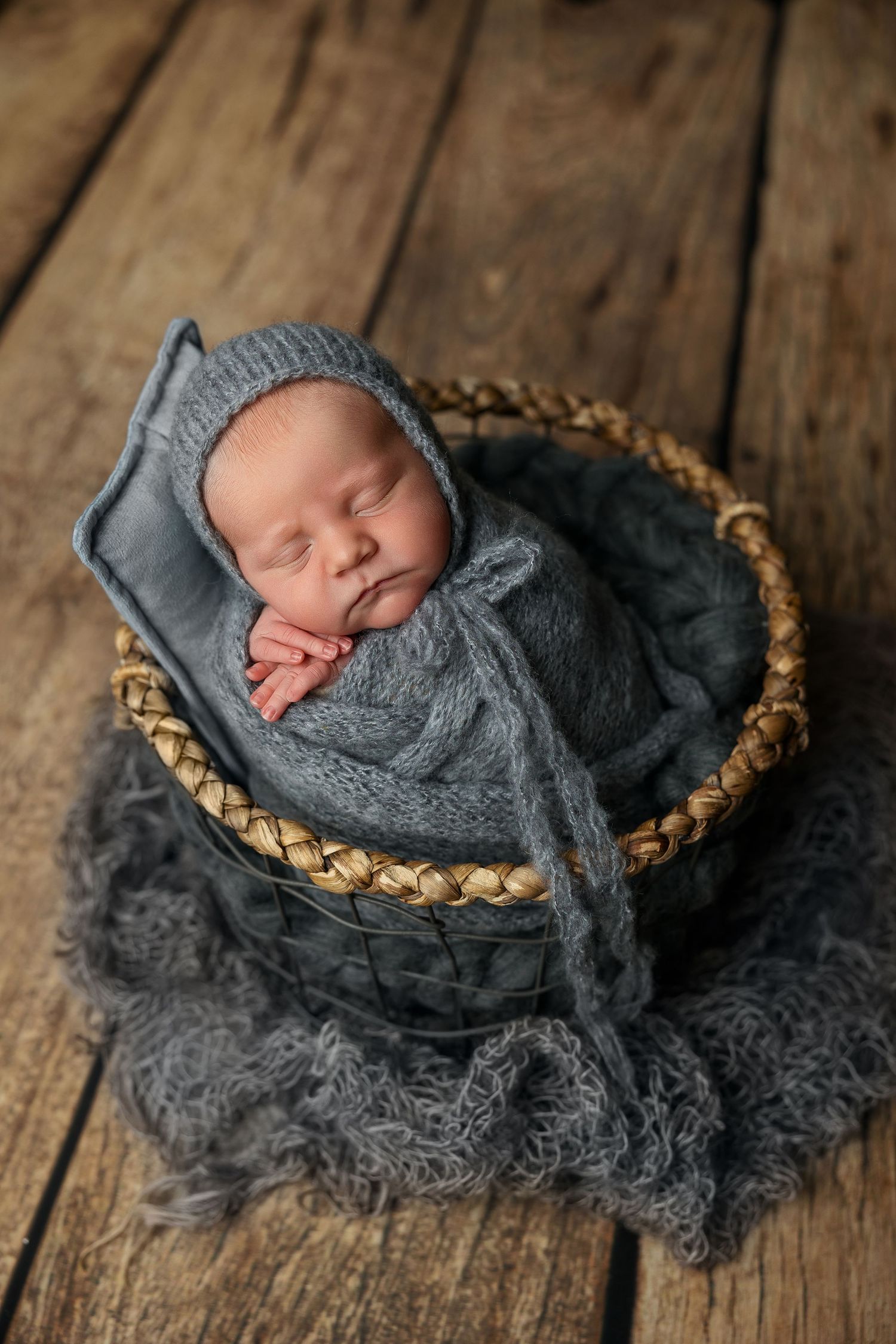 Newborn baby wrapped in soft gray blanket sleeps peacefully in woven basket on rustic wooden floor.