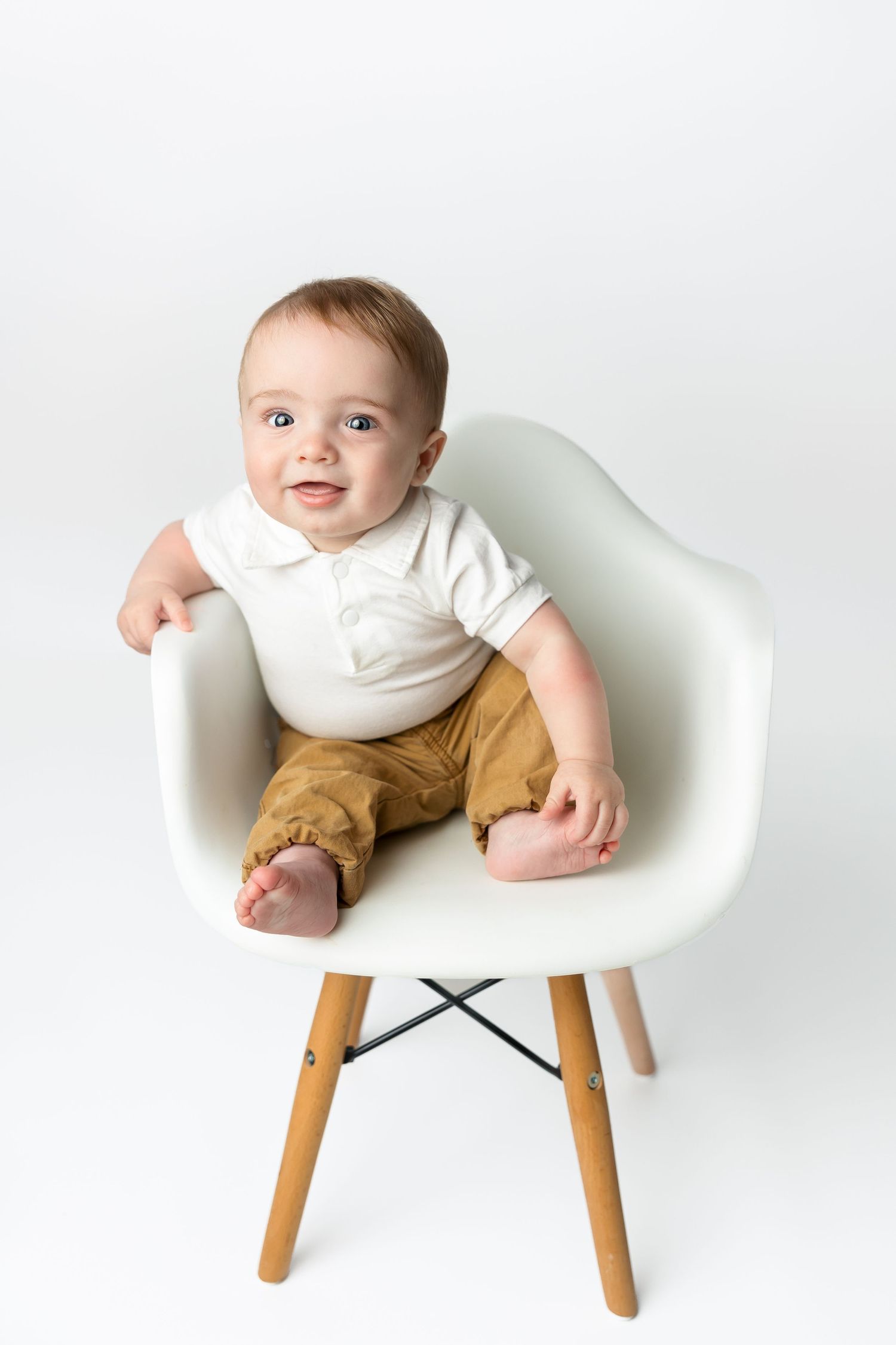 Baby wearing white shirt and brown pants sitting in modern white chair.